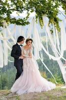 gli sposi si abbracciano sotto una vecchia quercia. servizio fotografico di matrimonio in montagna. accanto a loro viene preparato l'arredamento per la cerimonia. foto