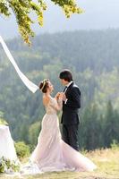 gli sposi si abbracciano sotto una vecchia quercia. servizio fotografico di matrimonio in montagna. accanto a loro viene preparato l'arredamento per la cerimonia. foto