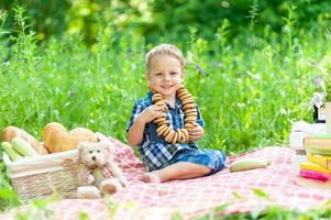 il ragazzino carino si siede su un plaid e si diverte in estate nella natura. sul collo del ragazzo c'è una collana di bagel.