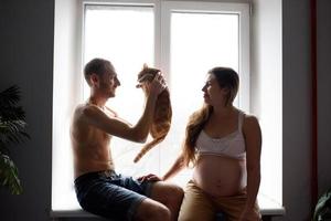 una coppia innamorata incinta è seduta sul davanzale della finestra e gioca con il loro gatto. autoisolamento di una casa durante la quarantena. foto