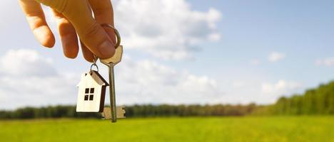 ciondolo in legno di una casa e chiave. sfondo del cielo e del campo. sognare casa, costruire un casolare in campagna, pianificare e progettare, consegna del progetto, trasloco in una nuova casa. copia spazio foto