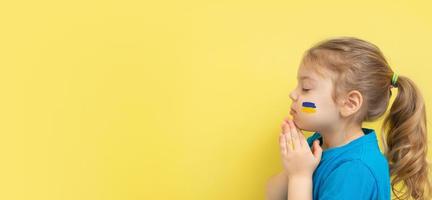 ragazza che tiene le mani giunte in preghiera con i colori giallo e blu della bandiera ucraina sulla guancia. copia spazio foto