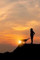 silhouette di lavoratore con carriola al tramonto, uomo agricoltore con carrello a spinta vuoto con tracciato di ritaglio foto