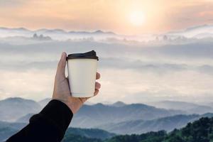mano maschile che tiene una tazza di carta da caffè calda con paesaggio di montagne sfocate sullo sfondo del mattino. foto