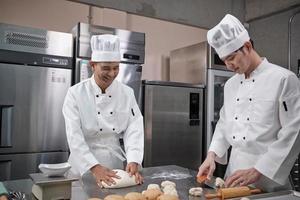 due cuochi professionisti asiatici maschi in uniformi bianche da cuoco e grembiuli stanno impastando pasta e uova, preparando pane e prodotti da forno freschi, cuocendo in forno nella cucina in acciaio inossidabile del ristorante. foto