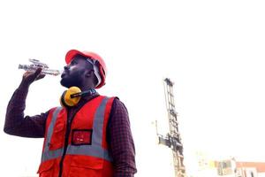 ritratto di un ingegnere lavoratore afroamericano che indossa un giubbotto e un casco di colore rosso neon luminoso di sicurezza, acqua potabile dalla bottiglia e in piedi al cantiere logistico del container del carico. foto