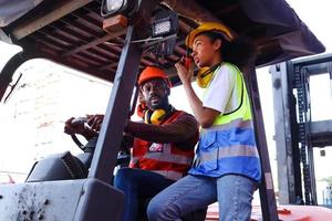 i lavoratori dell'industria indossano giubbotto di sicurezza e casco che guidano l'auto del carrello elevatore nell'industria della fabbrica di piante, due ingegneri afroamericani, un uomo e una donna, lavorano insieme nel cantiere di container logistici per il trasporto di merci. foto