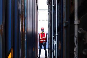 lavoratore ingegnere afroamericano che indossa un giubbotto e casco di colore rosso neon luminoso di sicurezza, con in mano una tavoletta digitale e in piedi tra i container nel deposito di container logistici per il trasporto di merci.