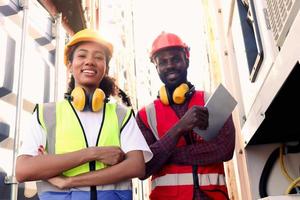 Ritratto di due uomini e donne sorridenti industriali afroamericani lavoratore ingegnere uomo e donna che indossano giubbotto di sicurezza e casco, in piedi con le braccia incrociate al cantiere logistico di container cargo. foto