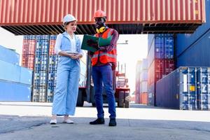 due lavoratori che indossano un giubbotto di sicurezza e un casco che discutono al cantiere di container per carichi logistici. uomo dell'ingegnere afroamericano che parla con il capo della bella giovane donna con i capelli biondi sul posto di lavoro. foto