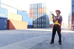 ingegnere lavoratore asiatico anziano che indossa giubbotto di sicurezza e casco, tiene in mano walkie-talkie radiofonici e indica qualcosa al cantiere logistico di container per carichi di spedizione. anziani al concetto di posto di lavoro foto