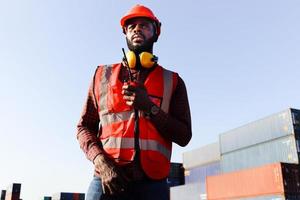 ritratto di un giovane ingegnere afroamericano lavoratore uomo che indossa un giubbotto e casco di sicurezza di colore rosso neon luminoso, parlando con un collega con walkie-talkie presso il cantiere logistico del container del carico di spedizione. foto