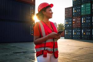 lavoratrice ingegnere donna afroamericana con capelli ricci che indossa giubbotto di sicurezza e casco, tenendo il telefono cellulare al tramonto al crepuscolo nel cantiere logistico del container del carico di spedizione. foto