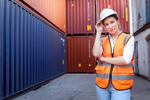 Ritratto di giovane e bella donna ingegnere capo sorridente felice con capelli biondi che indossa giubbotto di sicurezza e casco, in piedi con il braccio di fiducia incrociato al cantiere di container di carico logistico. foto