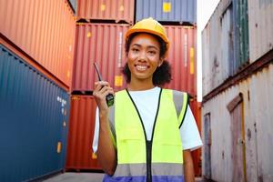 Ritratto di felice sorridente bella donna afroamericana ingegnere lavoratore con capelli ricci che indossa giubbotto di sicurezza e casco, tenendo walkies-talkie al cantiere di container di carico logistico. foto