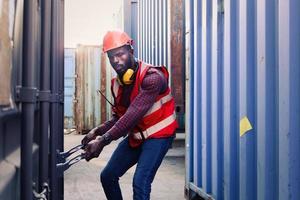 Ritratto di giovane ingegnere afroamericano lavoratore uomo che indossa un giubbotto e casco di sicurezza di colore rosso neon luminoso, cercando di aprire una porta del container di spedizione presso il cantiere di container logistici. foto