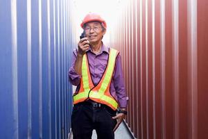 ritratto di ingegnere lavoratore asiatico anziano che indossa giubbotto di sicurezza e casco, con in mano walkie-talkie radiofonici, in piedi tra contenitori rossi e blu nel cantiere logistico di container cargo. foto