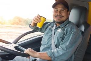 camionista asiatico uomo sorridente allegro in cabina di pilotaggio. fattorino seduto, bere caffè e rilassarsi. il camionista professionista ha fiducia e fiducia nel settore dei trasporti da molto tempo. foto