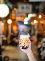 La giovane ragazza sta tenendo e mostrando una tazza di tè al latte con bolle di tapioca aromatizzato allo zucchero di canna nel mercato notturno di Taiwan sfondo, primo piano, bokeh foto