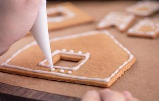 la donna sta decorando la casa dei biscotti di pan di zenzero con glassa bianca che guarnisce la crema di glassa sul fondo della tavola di legno, carta da forno in cucina, primo piano, macro. foto