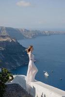 donna felice in abito bianco e cappello di paglia che si gode le vacanze sull'isola di santorini. vista sul mar egeo da oia. destinazione di viaggio estiva in europa. isole greche foto