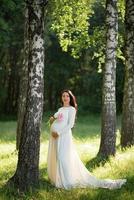 donna incinta in un cappello che posa in un vestito su uno sfondo di alberi verdi. foto