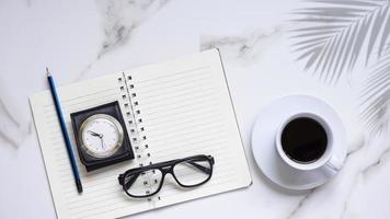 piatto di caffè nero in tazza di ceramica bianca, occhiali da vista, orologio da tasca vintage con custodia in pelle da viaggio, matita con blocco note a spirale e ombra di foglia di palma su piano in marmo bianco foto