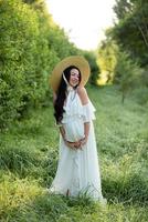 donna incinta in un cappello che posa in un vestito su uno sfondo di alberi verdi. foto