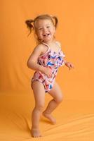 una bambina in costume da bagno all'età di un anno e mezzo salta o balla. la ragazza è molto felice. foto scattata in studio su sfondo giallo.