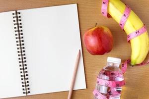 concetto di alimentazione sana, metro a nastro, frutta e bottiglia d'acqua su uno sfondo di legno, taccuino con spazio vuoto per la copia foto