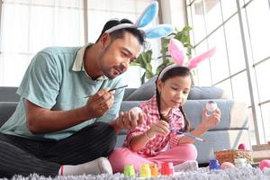 padre figlia prepara bellissime uova di Pasqua per la decorazione e la celebrazione dell'inizio della primavera, adorabile coniglietta rosa e suo padre con la fascia delle orecchie di coniglio che dipingono insieme uova colorate. foto