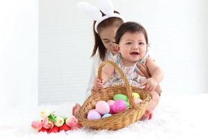 felice sorridente adorabile bambina di sei mesi che gioca con un cesto di vimini di uova di Pasqua colorate nell'abbraccio delle braccia della madre, mamma con la fascia delle orecchie di coniglio che tiene la sua dolce figlia piccola. foto