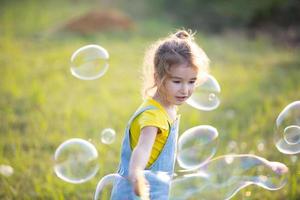 una ragazza con una tuta di jeans soffia bolle di sapone in estate in un campo al tramonto. giornata internazionale dei bambini, bambino felice, attività all'aperto. sfondo estivo. stile di vita sano ed ecologico foto