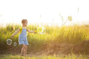 una ragazza con una tuta di jeans soffia bolle di sapone in estate in un campo al tramonto. giornata internazionale dei bambini, bambino felice, attività all'aperto. sfondo estivo. stile di vita sano ed ecologico foto