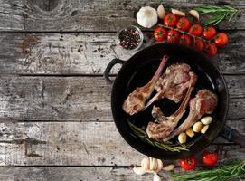 Costolette di agnello arrosto con rosmarino, pomodori e aglio in padella su sfondo di legno rustico vecchio scuro, vista dall'alto, spazio di copia foto