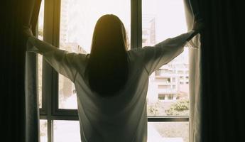 gioiosa giovane bella donna in piedi vicino alla camera da letto e tende aperte guardano in lontananza finestra incontrare benvenuto nuovo giorno. la donna sorridente si sente entusiasta delle opportunità o delle prospettive di carriera della vita. foto