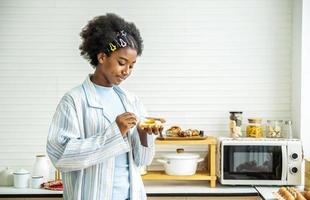 giovane donna afroamericana che sparge marmellata sul pane e mangia in cucina, lo studente gode della colazione o degli spuntini di pane tostato con marmellata foto