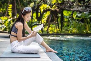 bella giovane donna asiatica si siede sul bordo della piscina all'aperto. lei legge un libro mentre si rilassa vicino alla piscina foto