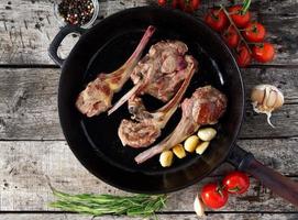 costolette di agnello arrosto con rosmarino, pomodori e aglio in padella su sfondo di legno rustico vecchio scuro, vista dall'alto foto