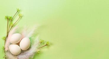 uova di Pasqua bianche in un delicato nido di piume colorate con boccioli di foglie spiegate sui rami. primavera, festa religiosa, nascita della vita. copia spazio. sfondo verde pallido foto