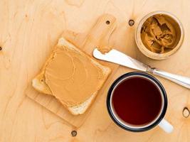 pane tostato con burro di arachidi, coltello da spalmare su un panino, una tazza di tè su fondo di legno beige foto