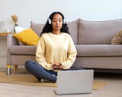 femmina afro-americana che ascolta la musica in cuffia mentre medita con il laptop a casa foto