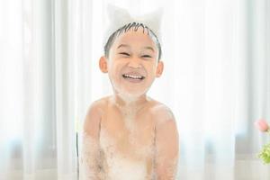 sorridente felice bambino asiatico ragazzo sta giocando con schiuma bianca nella vasca da bagno a casa. foto