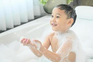 sorridente felice bambino asiatico ragazzo sta giocando con schiuma bianca nella vasca da bagno a casa. foto