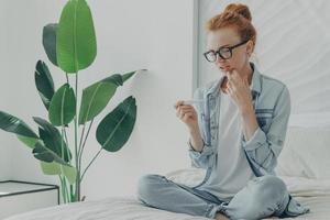 La giovane donna preoccupata dello zenzero controlla i risultati del test di gravidanza scopre la futura maternità si siede sul letto foto