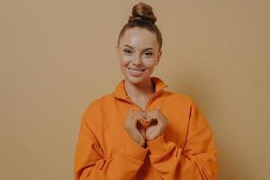 condividere l'amore. sorridente grata giovane donna che fa gesto del cuore, isolato su sfondo beige per studio foto