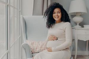 afro americana adorabile sorridente donna incinta in abito beige si siede in poltrona bianca foto