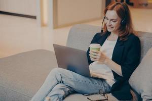 sorridente giovane agente del call center femminile in cuffia wireless e laptop seduto sul divano nella caffetteria foto