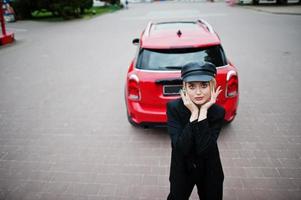 ritratto di bella modella bionda donna moda sexy in berretto e in tutto nero con trucco luminoso vicino a city car rossa. foto