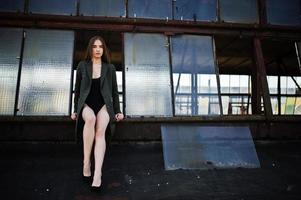 ragazza modello sexy con gambe lunghe in lingerie nera vestito corpo costume da bagno combidress e giacca poste sul tetto di un luogo industriale abbandonato con finestre. foto